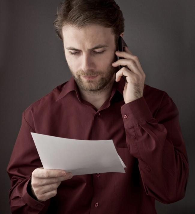Zdjęcie Forty years old office worker reading paper documents during mobile phone conversation &#8211; checking terms of agreement pochodzi z serwisu Shutterstock 